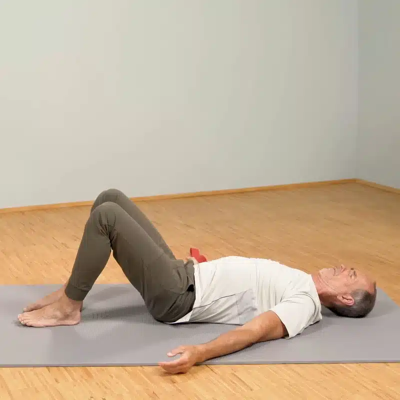 Roland Liebscher-Bracht liegt am Boden auf einer grauen Yoga-Matte. Seine Füße sind aufgestellt, während sein Rücken gerade am Boden liegt und er das Kinn nach unten zieht.