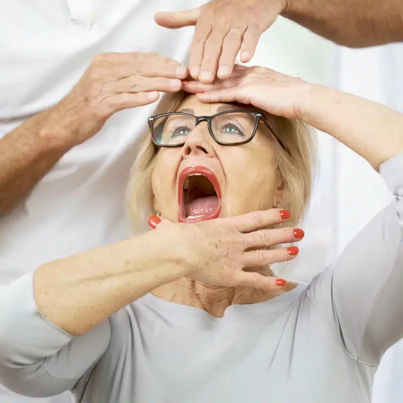 Eine Frau hat ihren Mund weit geöffnet. Mit einer Hand zieht sie ihren Unterkiefer herunter, während sie mit der anderen Hand ihren Kopf abstützt.