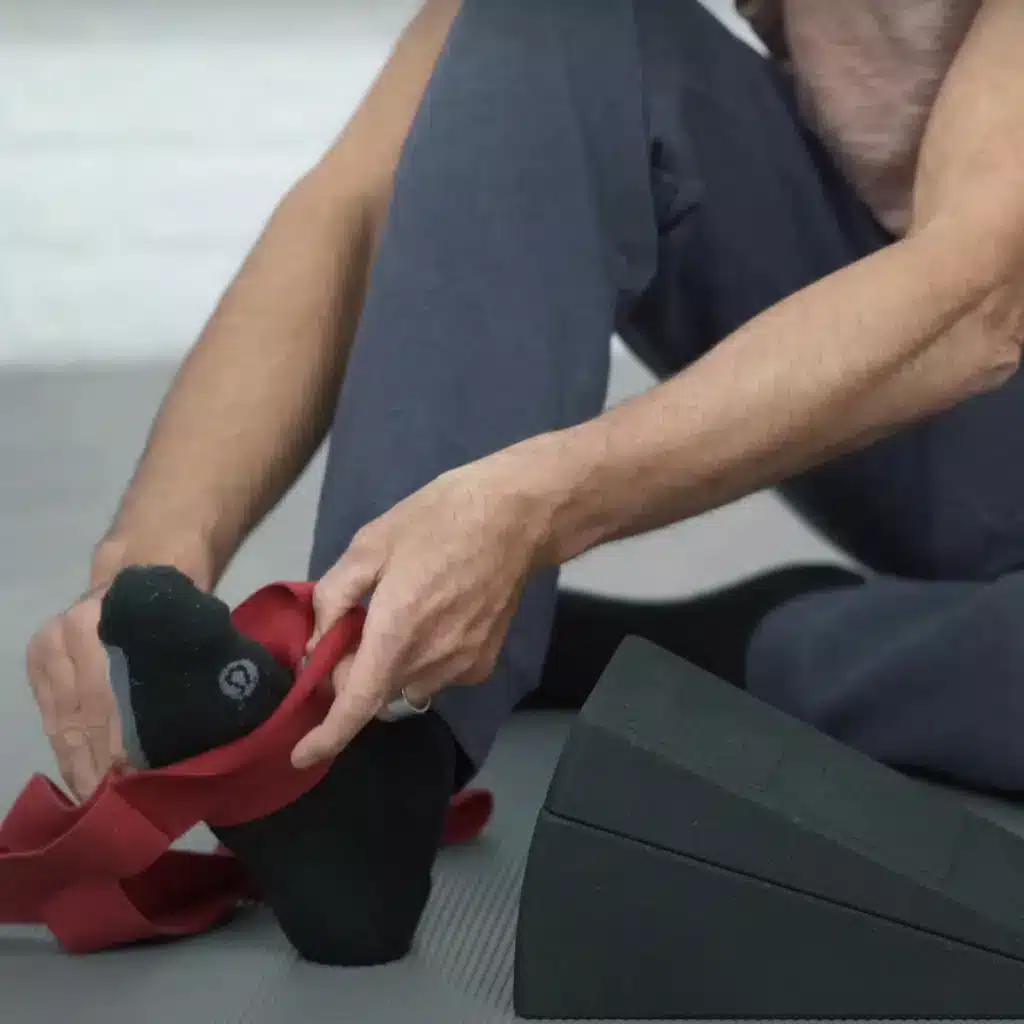 Eine Person sitzt auf dem Boden und fädelt den rechten Fuß in die Übungsschlaufe von Liebscher & Bracht ein.