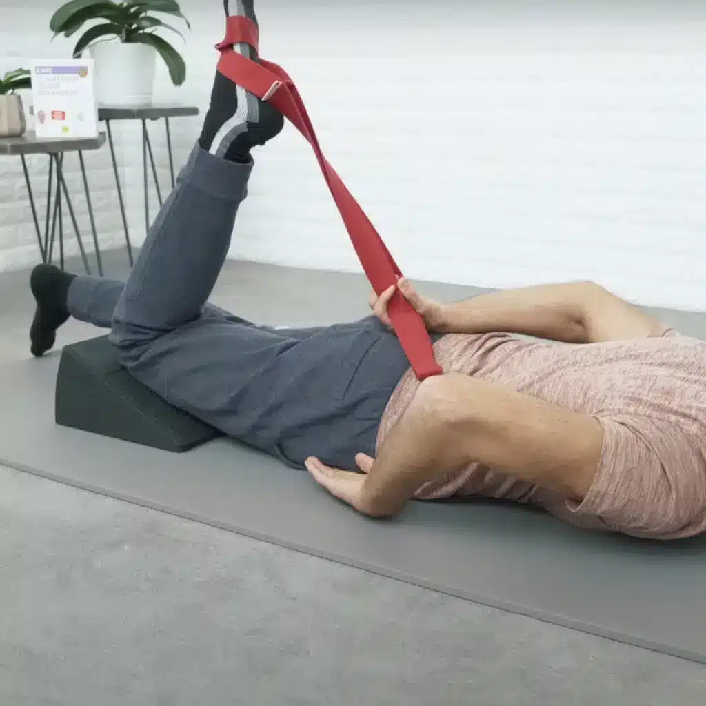 Eine Person liegt auf dem Bauch auf dem Boden und hat das rechte Knie angewinkelt. Der Fuß ist in die Übungsschlaufe eingefädelt und zieht in Richtung Gesäß.