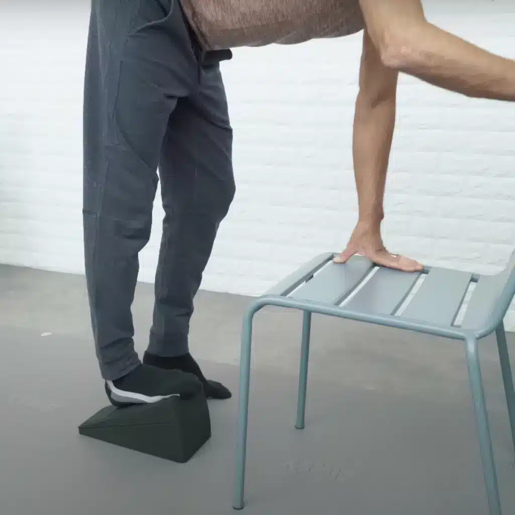 Eine Person steht vor einem Stuhl und stützt sich mit den Händen am Stuhl ab. Sie ist mit dem Oberkörper nach vorne geneigt und hat den rechten Fuß auf dem Knieretter abgestellt.