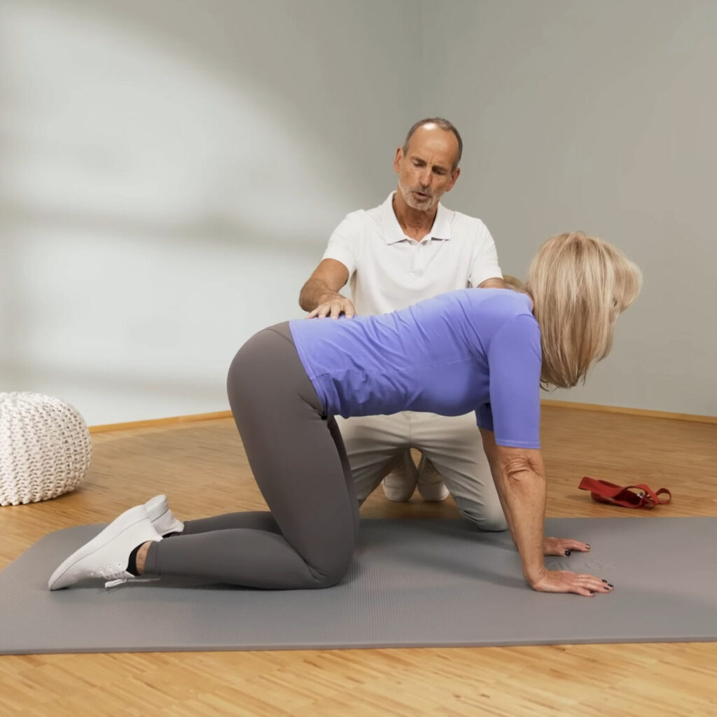 Eine Person steht auf allen vieren auf einer Matte. Den Rücken hält sie gerade und das Gesäß bewegt sie langsam zurück in Richtung Fersen.