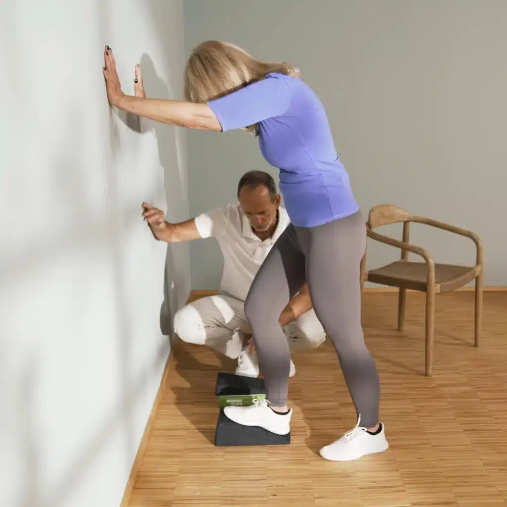 Eine Person steht vor einer Wand. Sie stützt sich mit beiden Händen an der Wand ab und steht mit dem rechten Fuß auf dem Knieretter von Liebscher und Bracht. Mit dem linken Fuß steht sie etwas weiter hinten.