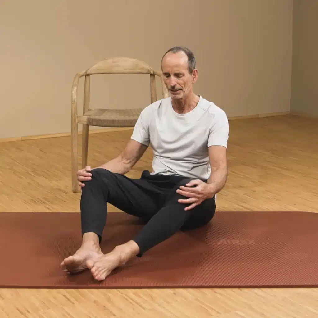 Eine Person sitzt auf einer Yogamatte und hat die Beine vor sich angewinkelt. Die Füße liegen aneinander und die Knie sind zu den Seiten geöffnet. Die Person umgreift die Knie mit den Händen.