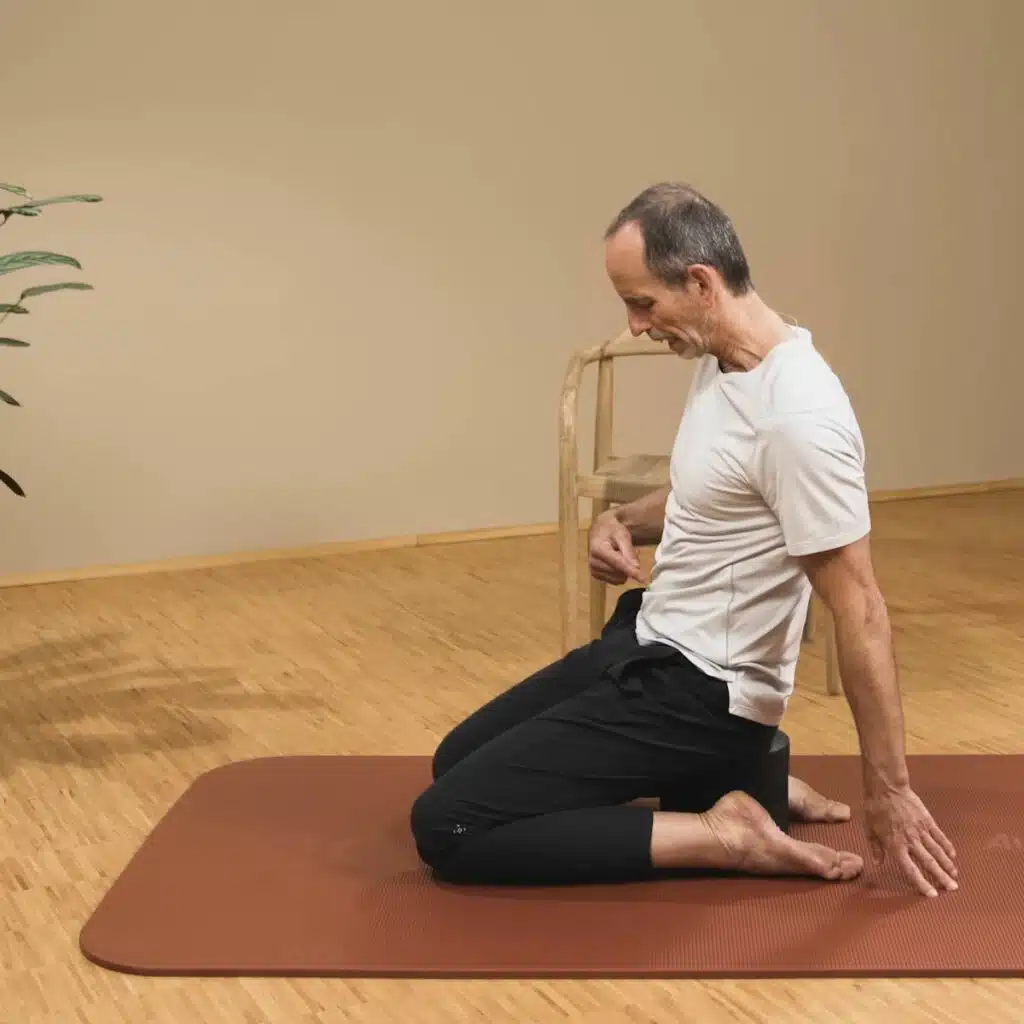 Eine Person sitzt auf dem Rückenretter auf einer Matte. Die Beine sind beide nach hinten angewinkelt. Außerdem zeigt die Person auf ihren Bauch, um zu zeigen, dass der Bauchnabel nach innen gezogen bleibt.