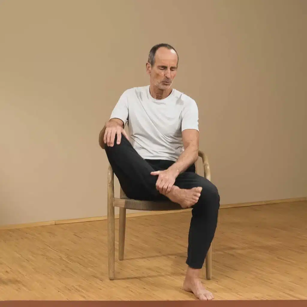 Eine Person sitzt auf einem Stuhl und zieht das angewinkelte, rechte Bein heran. Sie hält Knie und Sprunggelenk jeweils mit einer Hand fest.