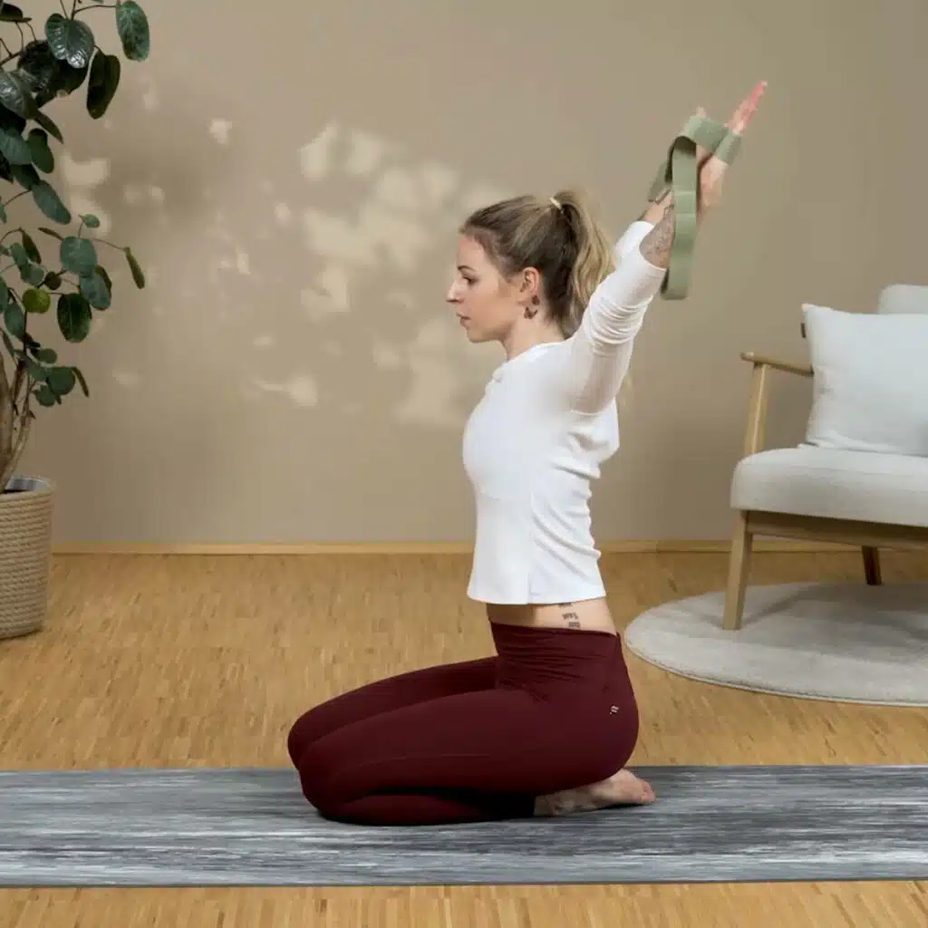 Eine Person hat beide Hände in die Schlaufen des Schulterretters gesteckt. Sie hält die gestreckten Arme weit geöffnet über den Kopf, sodass die Hände und die Arme sich hinter dem Kopf befinden. Sie kniet am Boden auf einer Matte.