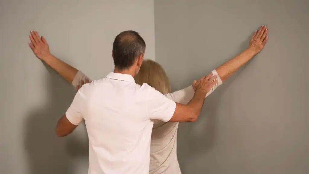 Eine Person steht in einer Raumecke und hat die Arme zu den Seiten ausgebreitet. Die Handflächen liegen an den Wänden und die Arme sind gestreckt.