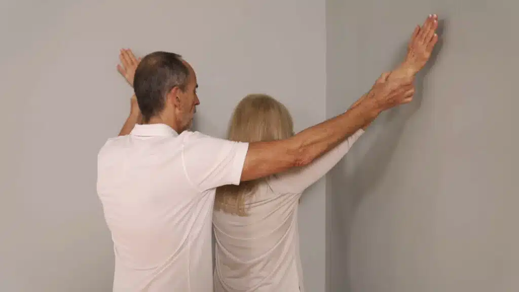 Eine Person steht in einer Raumecke und hat die Arme diagonal nach oben angehoben und die Handflächen an den Wänden abgelegt. Die Arme sind gestreckt.
