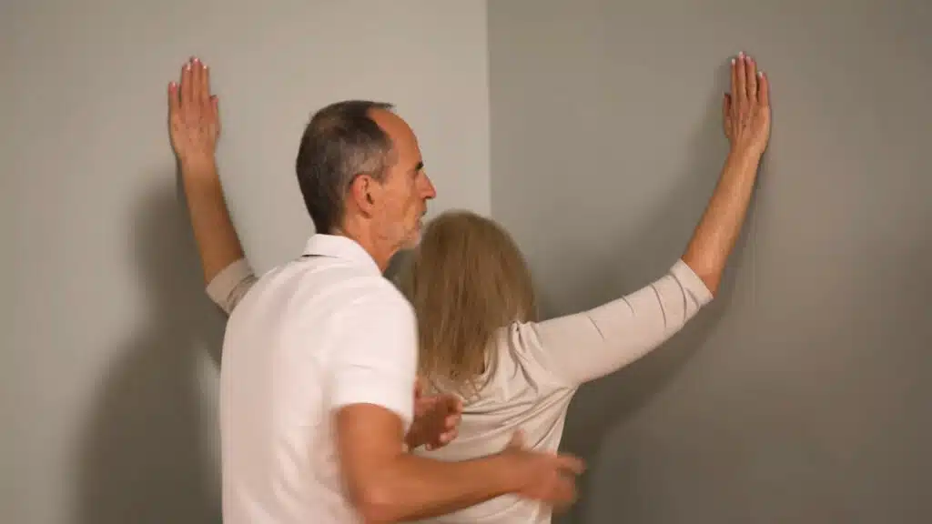 Eine Person steht in einer Raumecke und hat die Arme angewinkelt angehoben. Sie legt die Handflächen auf die Wände rechts und links der Ecke.