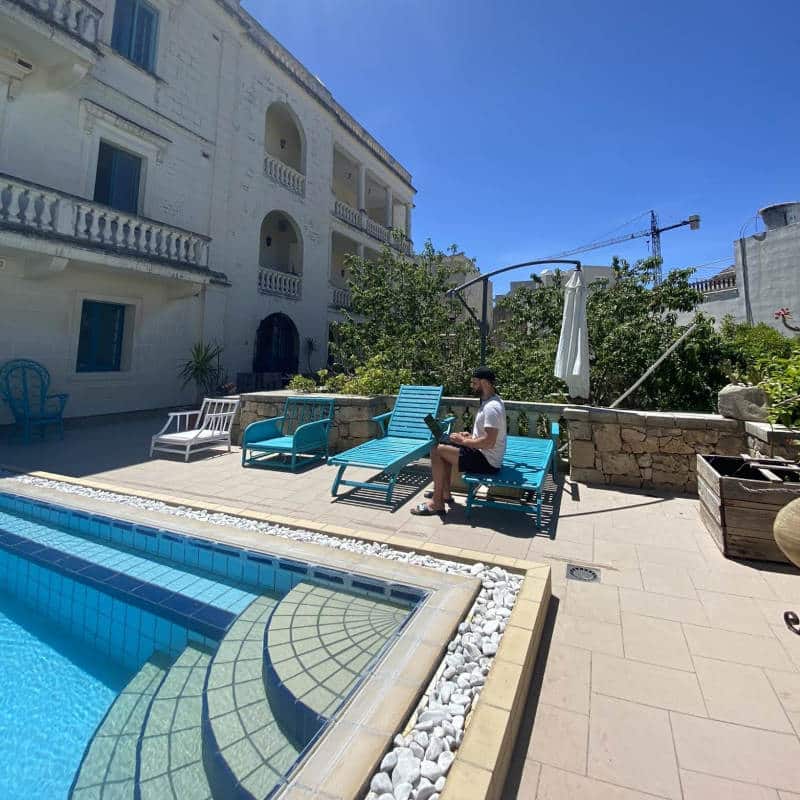 Der Eckbereich eines Swimming-Pools unter blauem Himmel, im Hintergrund ein junger Mann mit Laptop, der auf einer Liege sitzt