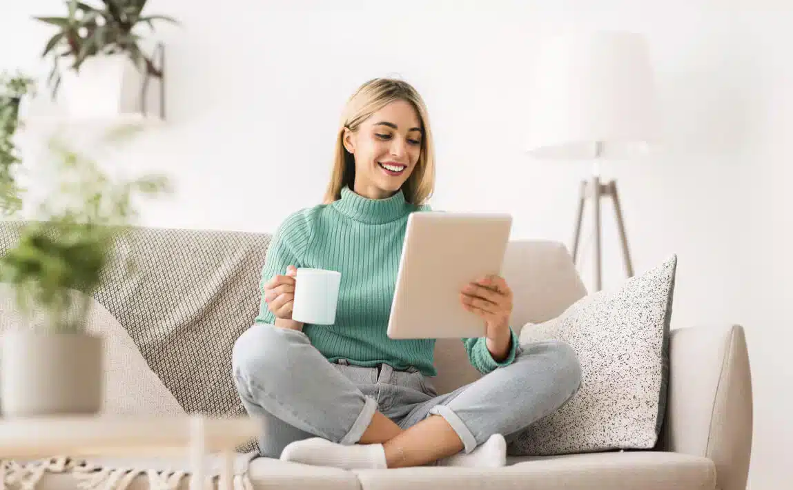 Eine Frau sitzt lächelnd im Schneidersitz auf einem Sofa und liest auf einem Tablet.