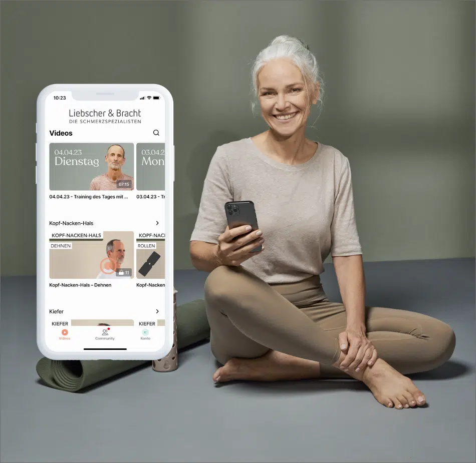 Eine ältere Frau mit grauen Haaren sitzt in beiger Sportkleidung auf dem Boden und hält ein Smartphone in der Hand. Neben ihr ist auf einem Smartphone die Liebscher & Bracht App eingeblendet.