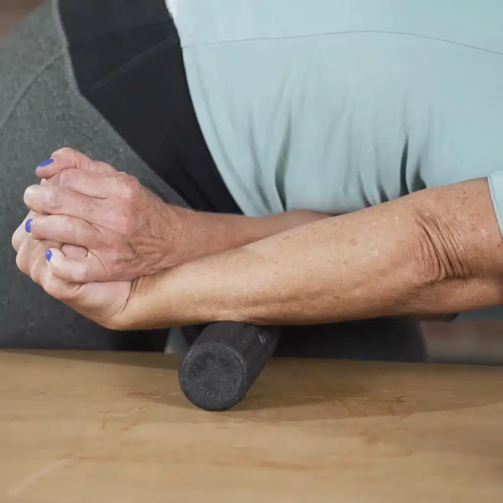 Hier sieht man eine Faszien-Rollmassage an der Oberseite des Unterarms. Dazu hat die ausführende Person eine Faust geballt und die Hand nach unten angewinkelt. Um den Arm mit der kleinen Faszienrolle zu massieren, hat die Person ihren Arm gestreckt und sich mit dem Oberkörper vorgebeugt, sodass die Oberseite des Unterarms auf der Rolle aufliegt.