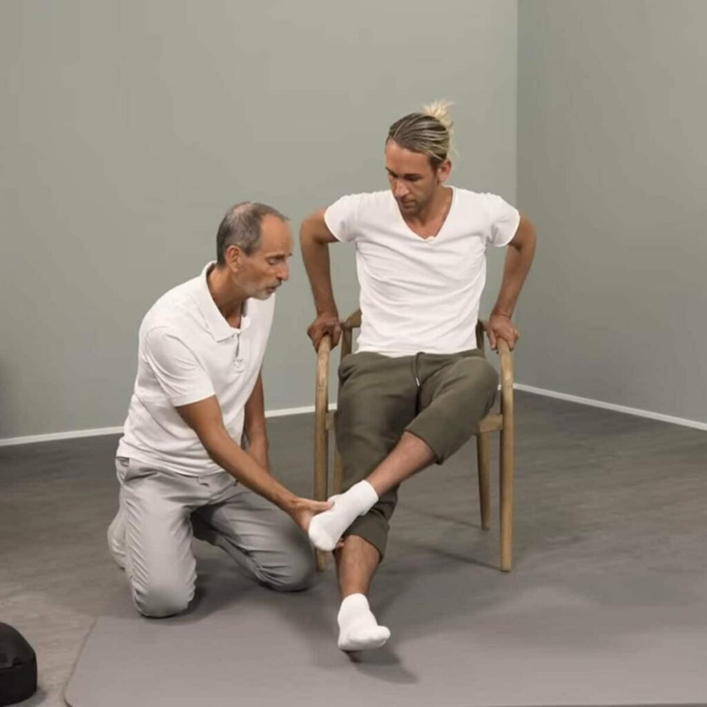 Eine Person sitzt auf einem Stuhl und hat das linke Bein leicht angewinkelt über das rechte überschlagen. Der linke Fuß befindet sich etwa beim rechten Schienbein.