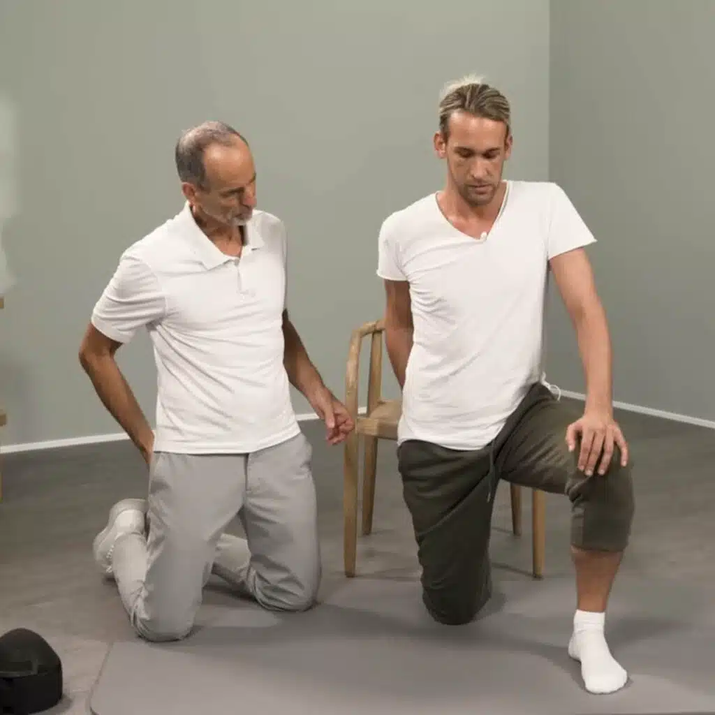 Eine Person kniet auf einer Übungsmatte und hat das rechte Knie am Boden, das linke Bein vorne aufgestellt. Die rechte Hand ist am rechten Gesäß und die linke Hand auf dem linken Knie. Der Oberkörper ist aufrecht