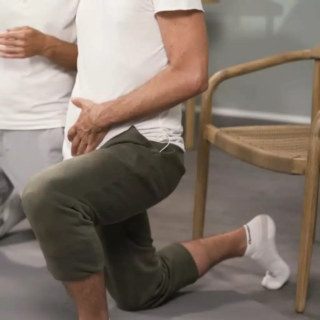 Eine Person kniet auf einer Übungsmatte und hat das rechte Knie am Boden, das linke Bein vorne aufgestellt. Die rechte Hand ist am rechten Gesäß und die linke Hand auf dem unteren Bauch. Der Oberkörper ist aufrecht.