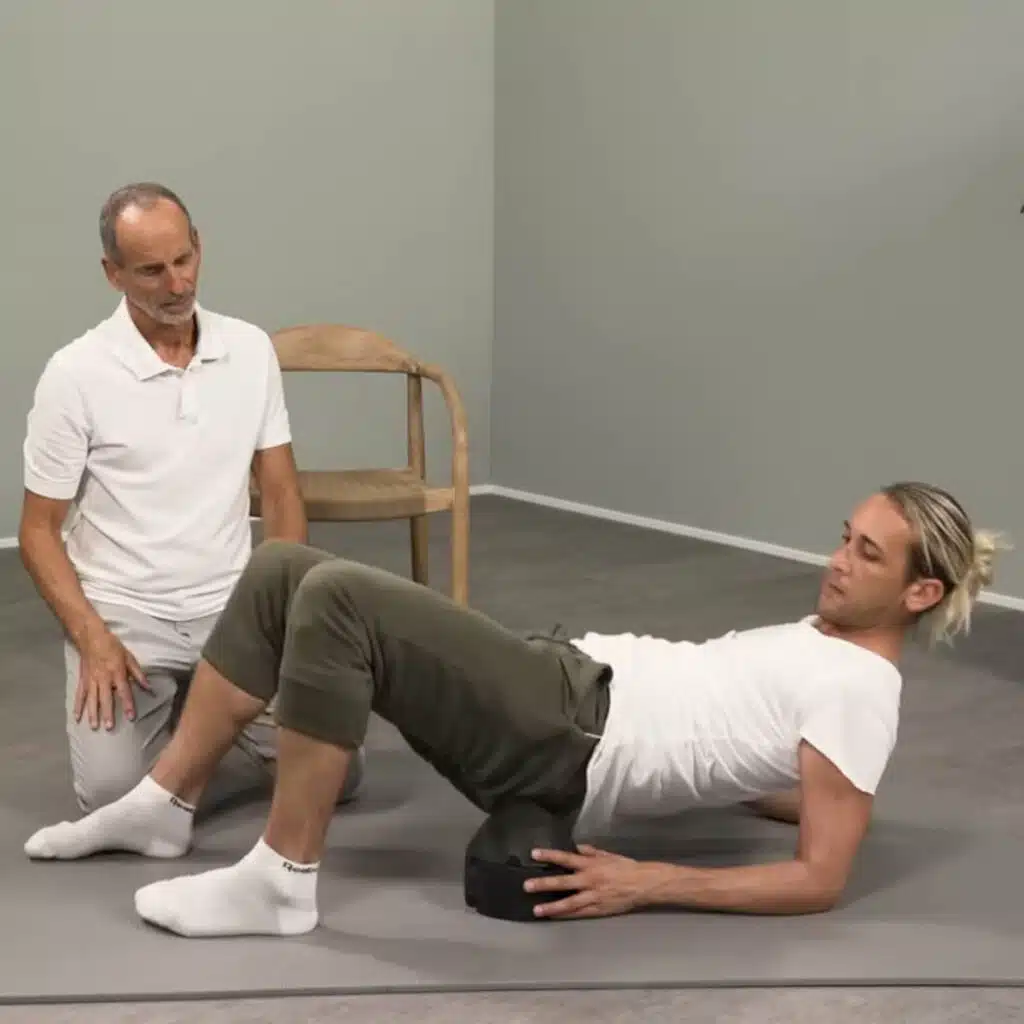 Eine Person liegt auf dem ISG-Ischias-Retter von Liebscher & Bracht. Sie hat den Retter auf einer Übungsmatte platziert, hält ihn mit einer Hand fest und legt das Gesäß vorsichtig drauf.