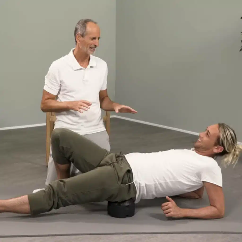 Eine Person liegt auf dem ISG-Ischias-Retter von Liebscher & Bracht. Sie hat den Retter auf einer Übungsmatte platziert und legt das Gesäß vorsichtig drauf. Mit den Unterarmen stützt sie sich am Boden ab.