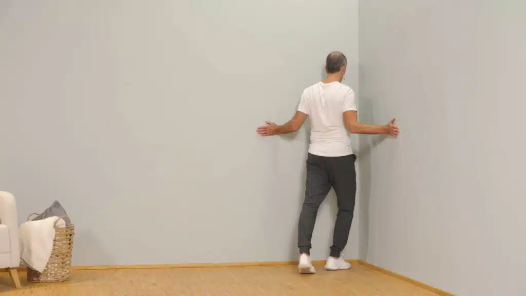 Eine Person steht vor einer Raumecke und hat die Arme zu den Seiten angewinkelt. Der Ellenbogen ist eng am Körper und die Arme sind in einer Außenrotation. So berühren die Handflächen die Wände rechts und links. Die Person macht einen Ausfallschritt und drückt den Brustkrb in die Raumecke.
