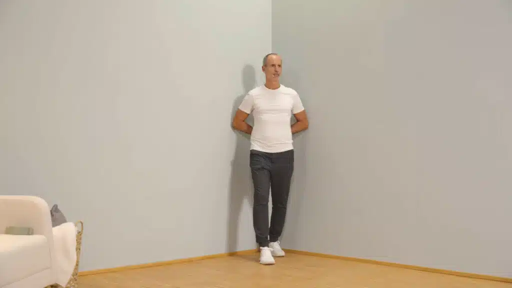 Eine Person steht mit dem Rücken zur Raumecke. Die Handrücken liegen auf dem unteren Rücken ab, die Arme sind angewinkelt. So befinden sich beide Arme in einer Innenrotation. Die Ellenbogen berühren rechts und links die Wände. Die Person lehnt sich mit dem Rücken in die Ecke, um die Schultern in der Innenrotation zu dehnen.
