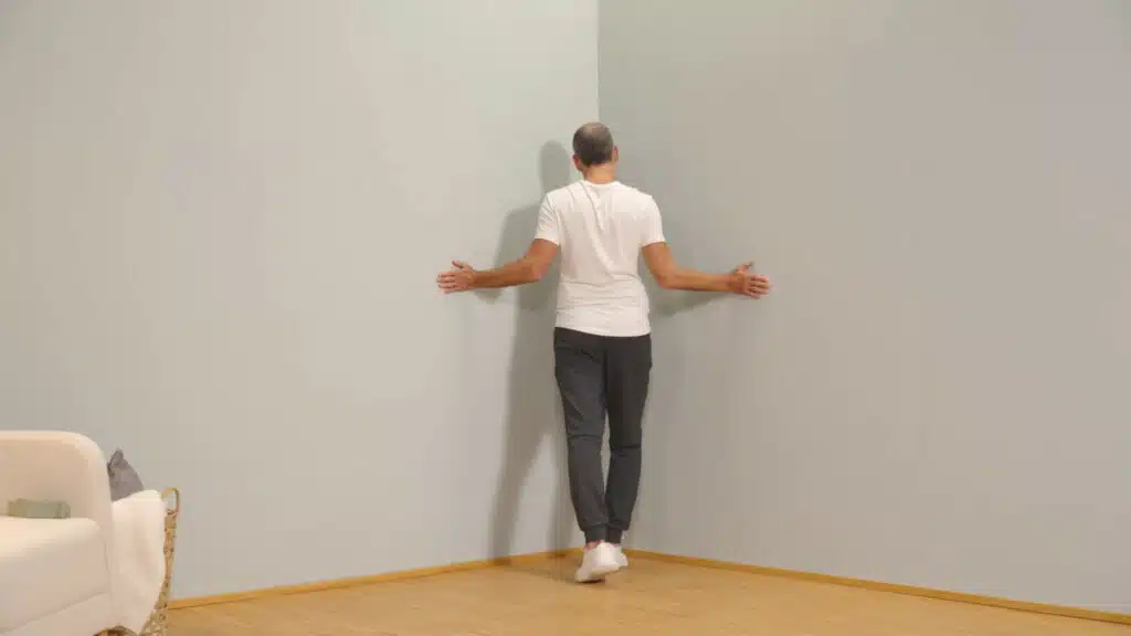 Eine Person steht vor einer Raumecke und hat die Arme zu den Seiten angewinkelt. Der Ellenbogen ist eng am Körper und die Arme sind in einer Außenrotation. So berühren die Handflächen die Wände rechts und links. Die Person macht einen Ausfallschritt und drückt den Brustkrb in die Raumecke.