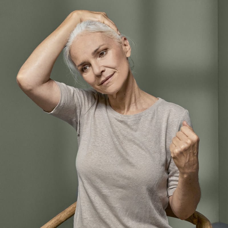 Eine Frau zeigt eine Übung gegen Nackenschmerzen. Ihr linker Arm ist gebeugt, die Hand zu einer Faust geschlossen. Die rechte Hand zieht den Kopf nach rechts.
