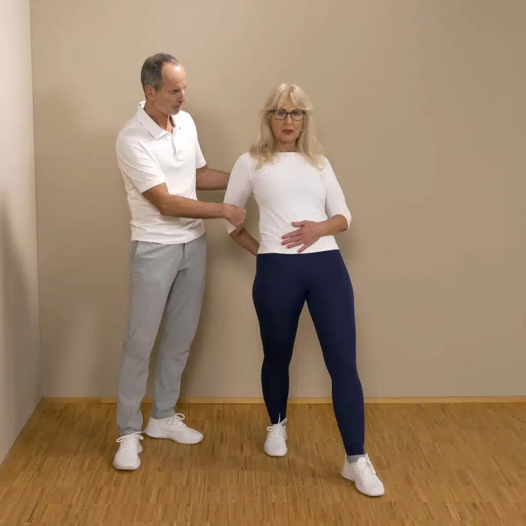 Eine Person macht einen Ausfallschritt mit rechts nach hinten. Sie streckt die Beine und hält die rechte Hand auf dem rechten Gesäß. Die linke Hand liegt auf dem unteren Bauch.