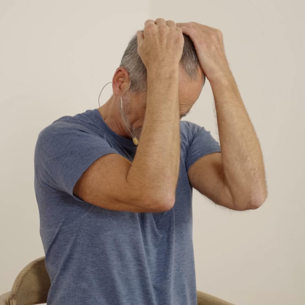 Roland Liebscher-Bracht sitzt auf einem Stuhl und hat beide Hände um den Kopf gelegt. Die Fingerspitzen berühren seinen Hinterkopf und er zieht den Kopf nach vorne unten, sodass sein Kinn in Richtung Brustkorb sinkt.