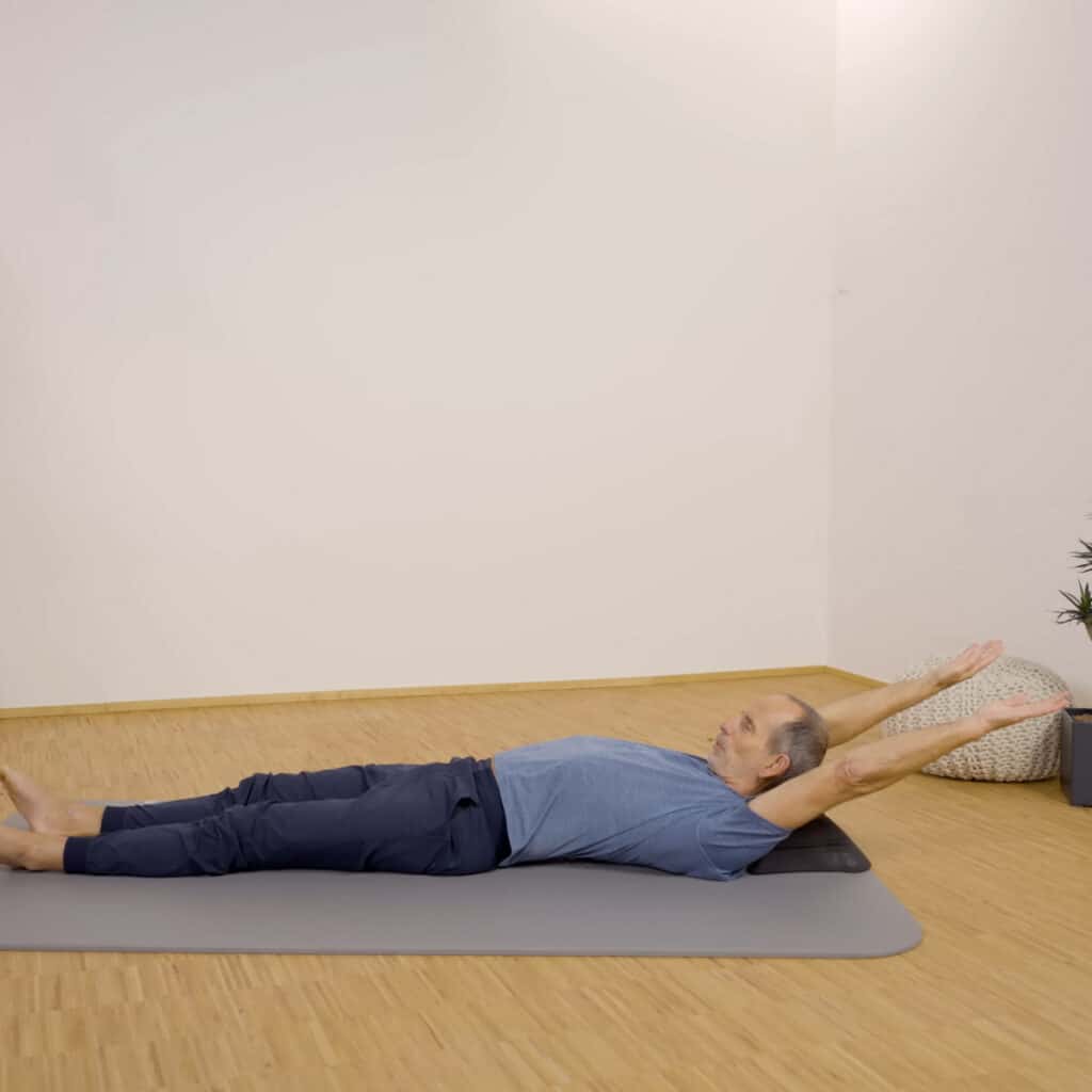 Roland Liebscher-Bracht liegt auf einer Übungsmatte auf dem Nackenretter und hat die Arme weit über den Kopf nach hinten gestreckt.