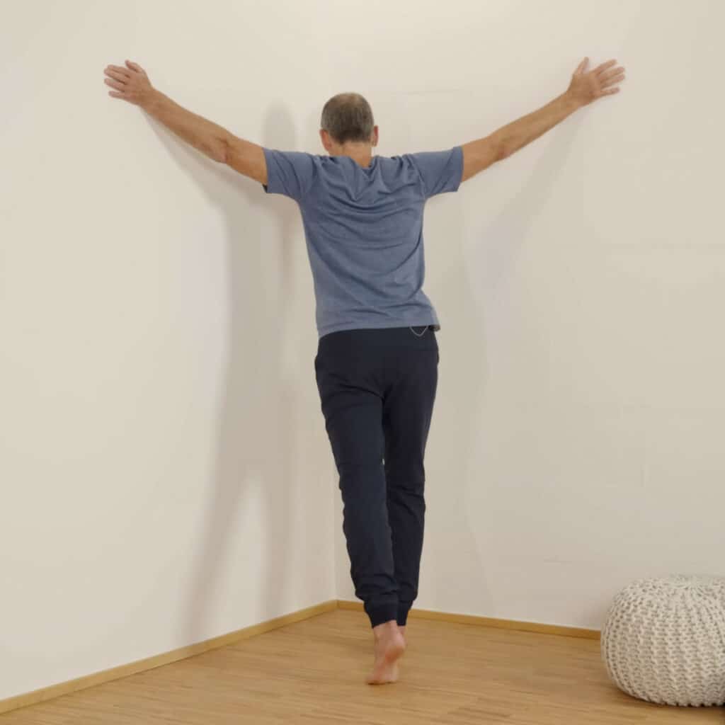 Roland Liebscher-Bracht steht in einer Raumecke und hat die Arme zu den Seiten ausgestreckt. Die Handflächen liegen auf den Wänden rechts und links ab und mit einem Ausfallschritt drückt er sein Brustbein in Richtung Ecke.