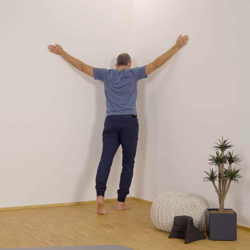 Roland Liebscher-Bracht steht in einer Raumecke und hat die Arme zu den Seiten ausgestreckt. Die Handflächen liegen auf den Wänden rechts und links ab und mit einem Ausfallschritt drückt er sein Brustbein in Richtung Ecke.