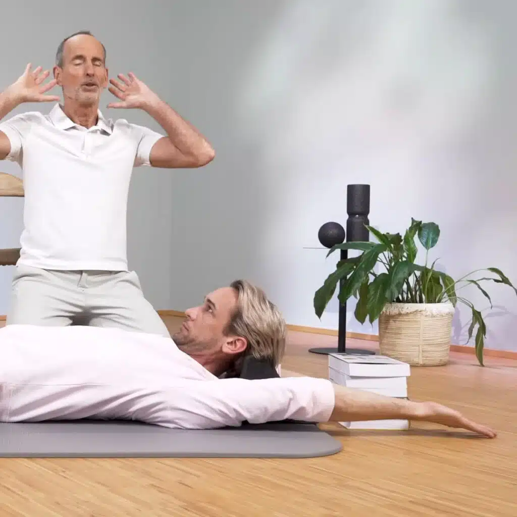 Eine Person liegt am Boden und hat den Kopf auf dem Nackenretter abgelegt. Das Kinn zieht zum Brustkorb. Zusätzlich streckt sie die Arme lang nach hinten aus.