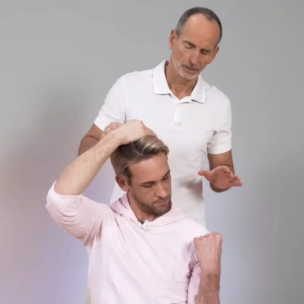 Eine Person hat den linken Arm angewinkelt und die linke Faust an der linken Schulter abgelegt. Der linke Ellenbogen ist eng am Körper. Sie schaut ihre Faust an. Die rechte Hand greift über den Kopf und zieht den Kopf nach rechts.