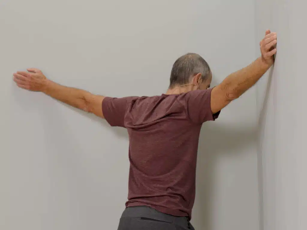 In einer Raumecke steht ein Mann und hat seine Arme zu beiden Seiten ausgestreckt, sodass seine Hände an den Wänden aufliegen und er sich in die Raumecke hineinlehnen und dehnen kann.