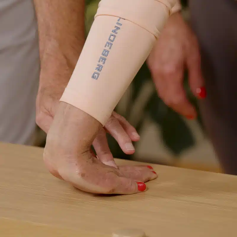 Eine Person hat ihre Handfläche auf einem Tisch abgelegt. Die Finger zeigen in Richtung ihrer Beine, das Handgelenk ist leicht vom Tisch angehoben.