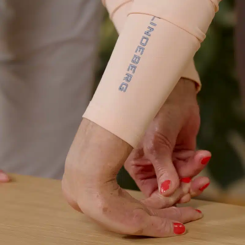 Eine Person hat ihre Handfläche auf einem Tisch abgelegt. Die Finger zeigen in Richtung ihrer Beine, das Handgelenk ist leicht vom Tisch angehoben. Die Person hat zusätzlich mit der anderen Hand den gedehnten Mittelfinger leicht angehoben.