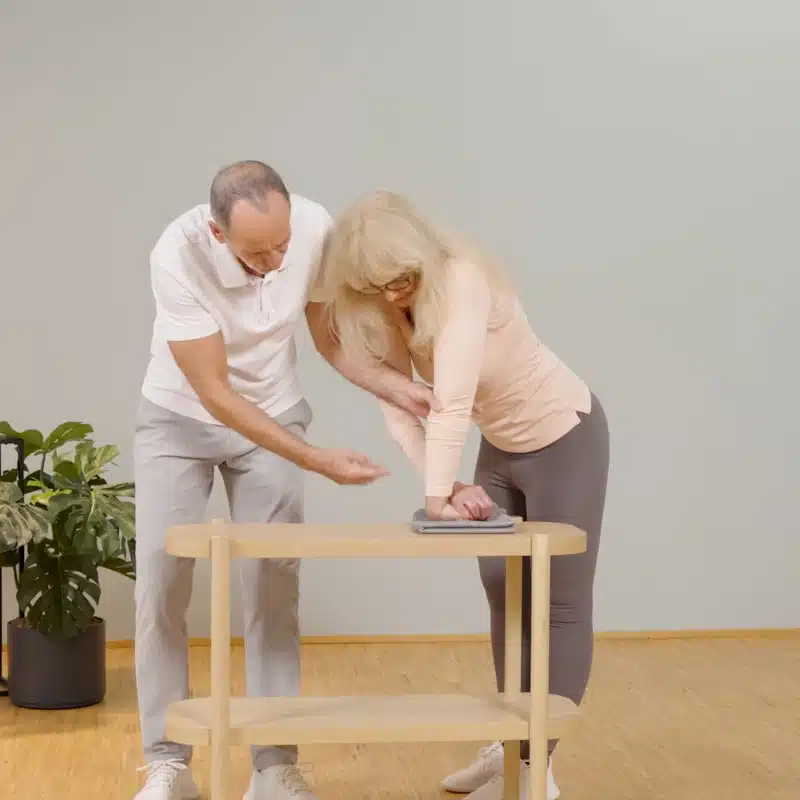 Eine Person hat ihre Hand zu einer Faust geballt, und den Handrücken auf einem Tuch auf einem Tisch abgelegt. Sie umgreift die Faust mit der anderen Hand und drückt sie nach unten. Der Arm der geballten Faust bleibt gestreckt.