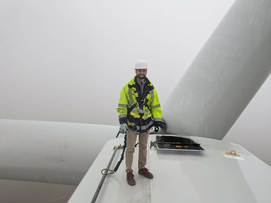 Man sieht einen Mann in gelber Arbeitsjacke und mit Helm auf dem Kopf oben auf einem Windkraftrad stehen und frontal in die Kamera lächeln.