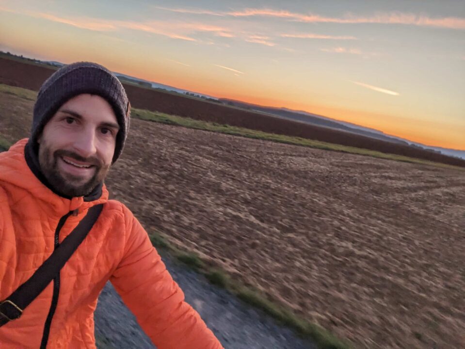 Man sieht im linken Bildteil einen lächelnden Mann im 3/4-Profil, der eine Mütze trägt vor einer Strandkulisse am Abend