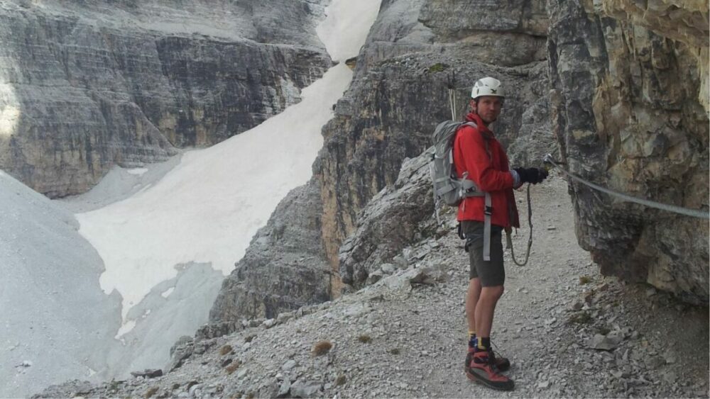 Ein Mann steht auf einem Weg, schaut über die rechte Schulter in die Kamera und ist mit Seilen an eine Felswand gesichert.