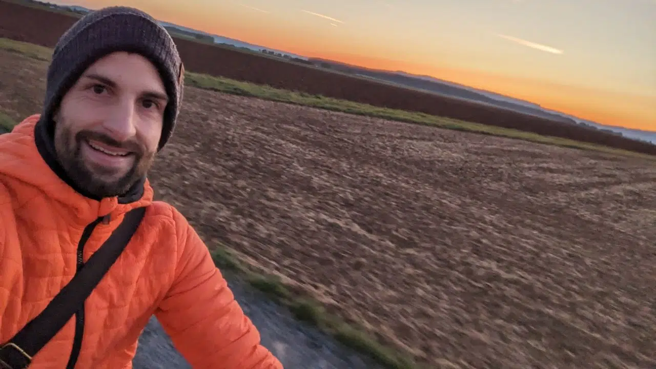 Im Hintergrund sieht man Felder, im Vordergrund links ist der Oberkörper und Kopf eines Mannes in einer orangenen Jacke, mit Mütze und Bart zu sehen, der in die Kamera lächelt