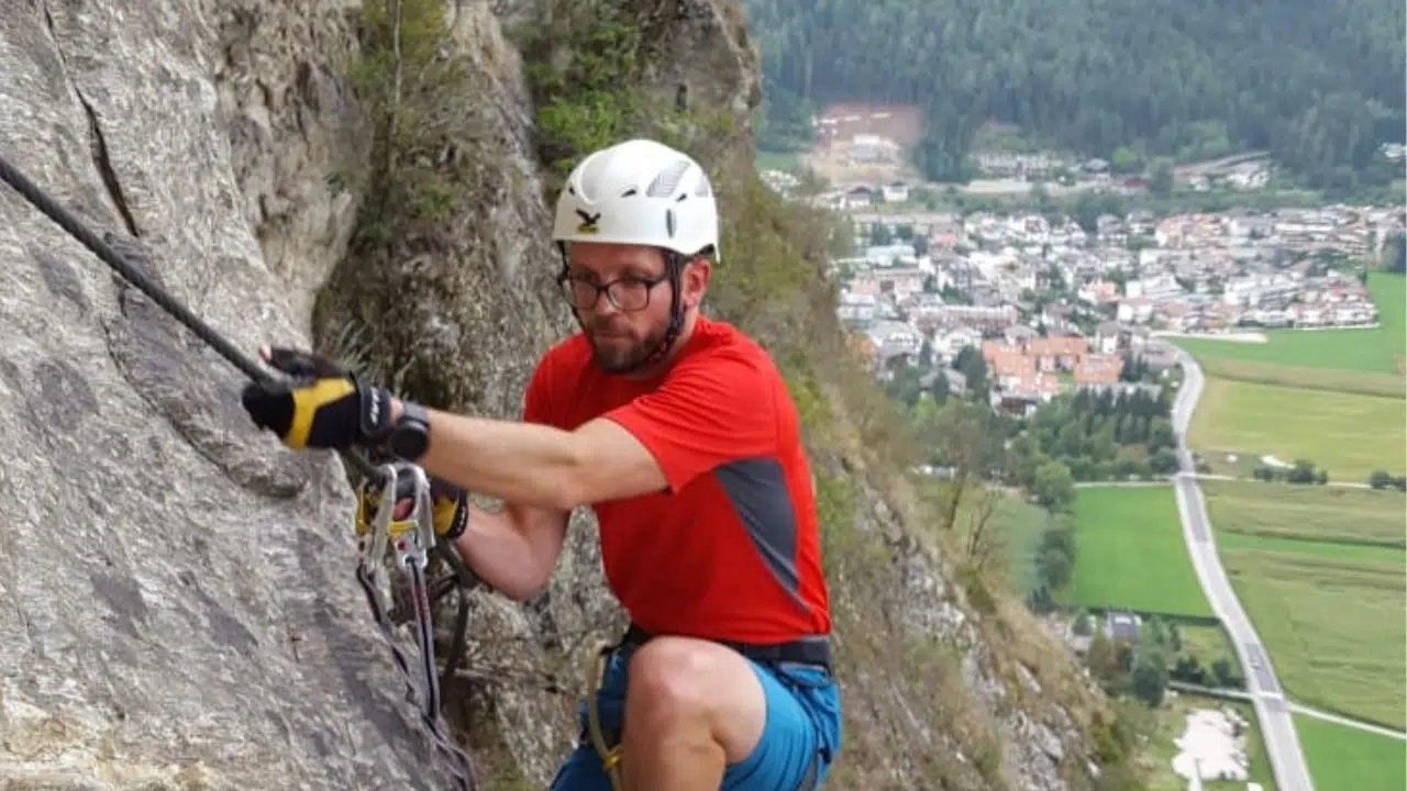 Ein Mann ist im Vordergrund an einer Kletterwand in den Bergen zu sehen, im Hintergrund sieht man ein Tal mit Häusern und einer Straße