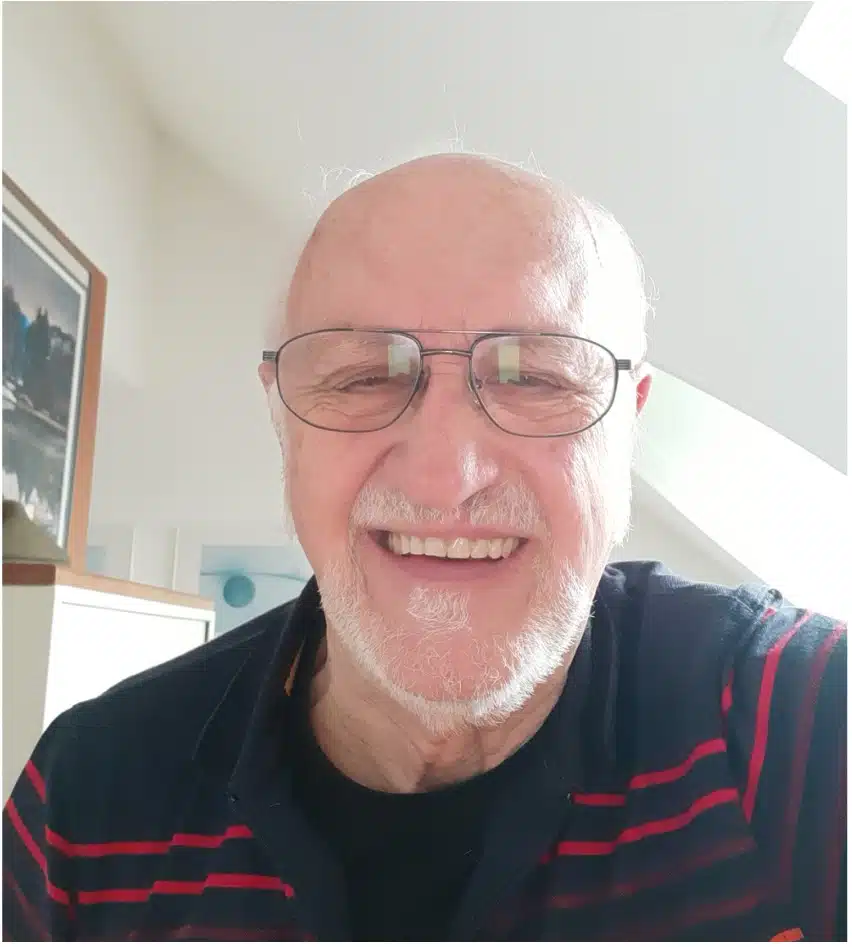 Man sieht einen weißhaarigen Mann mit Glatze, Bart und ovaler Brille frontal in die Kamera lächeln, im Hintergrund sieht man eine Dachschräge