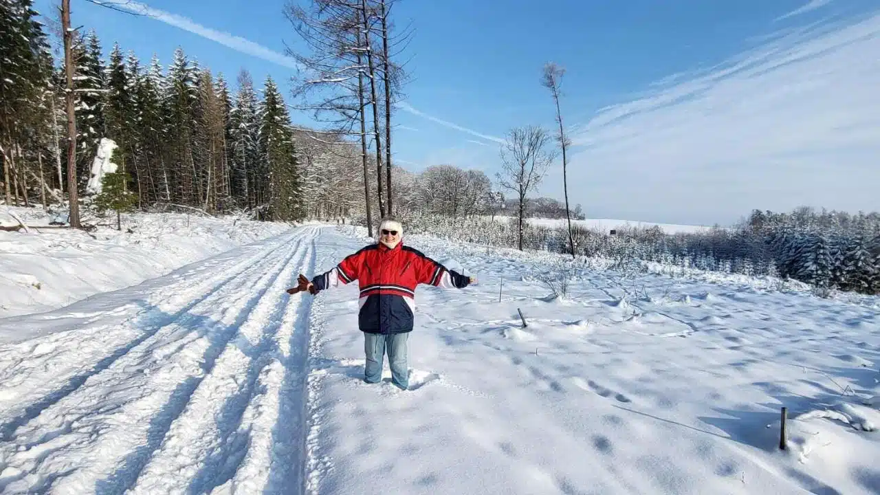 Winterlandschaft,eine Frau in einer roten Jacke steht mit ausgebreiteten Armen im Schnee und lächelt in die Kamera